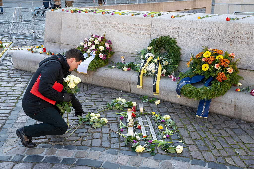 Ein Mann legt weiße Rosen nieder, dahinter liegen Blumenkränze an einer Gedenkstätte