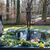 Zwei Personen stehen an einer Gedenkstätte mit einer Bronze-Skulptur, rundherum liegen Blumen