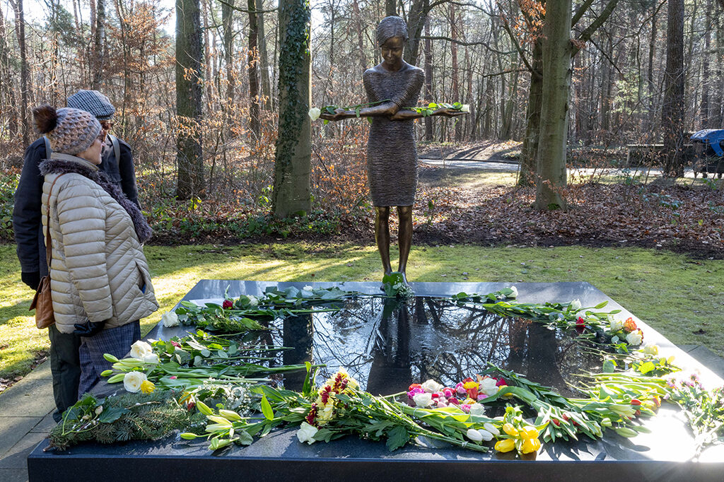Zwei Personen stehen an einer Gedenkstätte mit einer Bronze-Skulptur, rundherum liegen Blumen