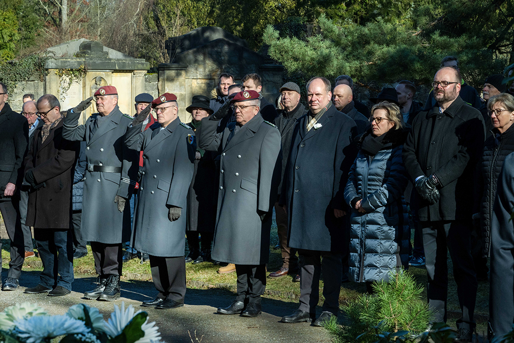Mehrere Personen stehen auf einem Friedhof zusammen, drei Mänenr tragen Militär-Uniform und salutieren