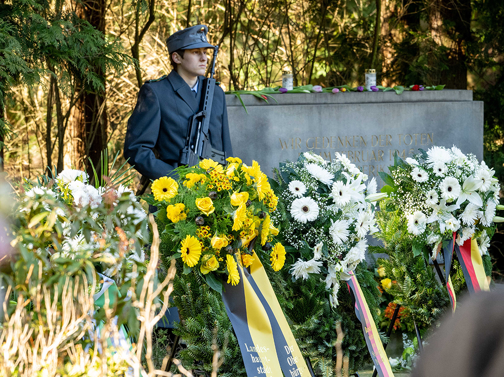 Blumengebinde stehen vor einer Gedenkstätte, daneben steht ein junger Mann in Militär-Uniform