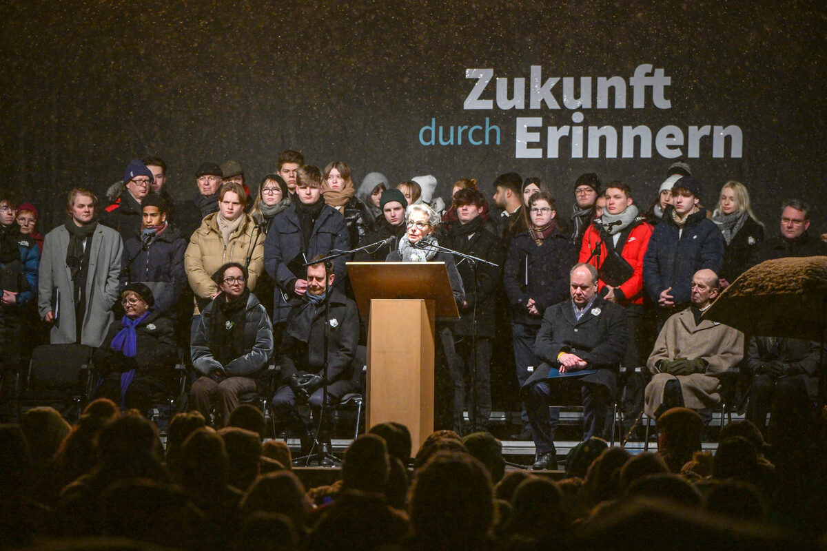 Mehrere Menschen stehen und sitzen auf einer Bühne. Die Rektorin der TU Dresden steht hinter einem Rednerpult.