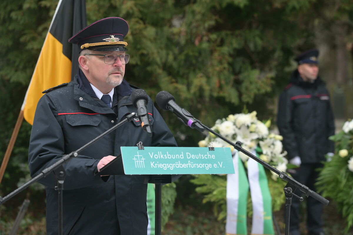 Ein Mann in Uniform steht an einem Rednerpult, im Hintergrund stehen Kränze